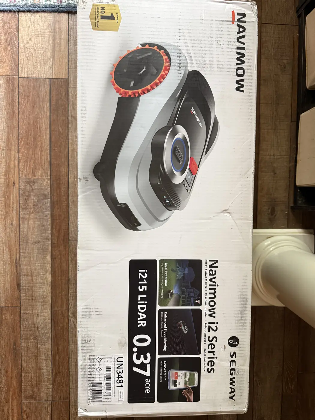 Sealed Segway Navimow i215 LiDAR retail box photographed on a hardwood floor, printed 'Navimow i2 Series, i215 LiDAR, 0.37 acre' with Dual Precision, Enhanced Slope Mowing and GeoSketch feature panels and a UN3481 lithium battery shipping label